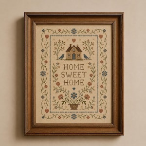 May include: A framed cross-stitch embroidery on beige fabric featuring the text "HOME SWEET HOME" in brown letters. The design includes a small house, two birds, a flower basket, and floral vines in shades of blue, brown, and red, all enclosed in a dark brown wooden frame.