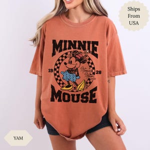 May include: A rust-coloured t-shirt with a Minnie Mouse graphic. The design includes the text "MINNIE MOUSE" in black, with the years "1928" and "xoxo". Minnie Mouse is shown in a blue dress and yellow shoes, within a circular design.