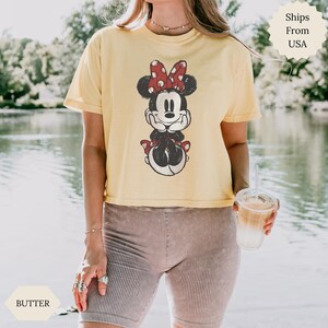 May include: A yellow t-shirt featuring a distressed Minnie Mouse graphic with a red bow. The shirt is paired with ribbed shorts. The image is taken outdoors near a body of water.