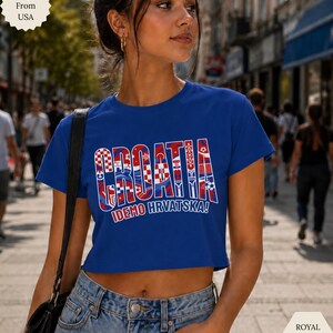 May include: A royal blue short-sleeved crop top featuring the word CROATIA in large block letters filled with red and white checkered patterns and soccer ball graphics, with the text IDEMO HRVATSKA! printed in white underneath the main design.