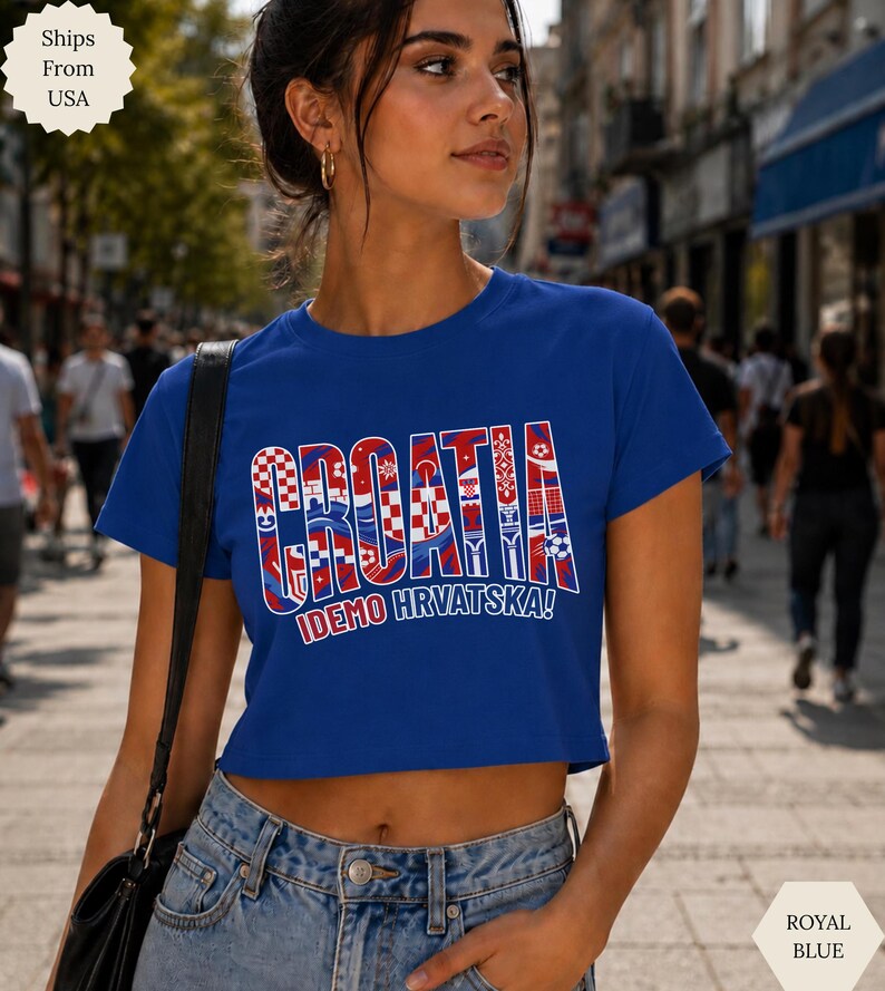 May include: A royal blue short-sleeved crop top featuring the word CROATIA in large block letters filled with red and white checkered patterns and soccer ball graphics, with the text IDEMO HRVATSKA! printed in white underneath the main design.