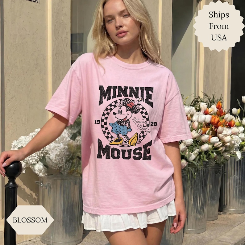 May include: A light pink t-shirt with a graphic of Minnie Mouse in a checkered circle. The words "MINNIE MOUSE" are above and below the image. The shirt has short sleeves and a loose fit.