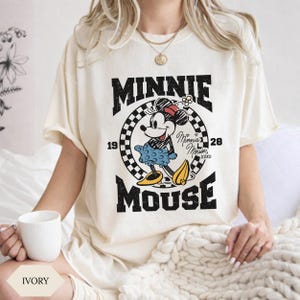 May include: Cream-coloured t-shirt with a vintage Minnie Mouse graphic. The design features the words "MINNIE MOUSE" in bold black letters, a chequered circle, and the years "19" and "28". The shirt has short sleeves.