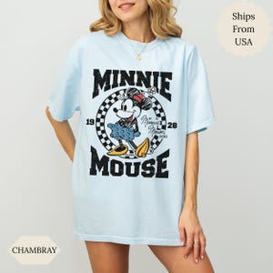 May include: Pale blue t-shirt with a distressed Minnie Mouse graphic. The design features the text "MINNIE MOUSE" in black, with Minnie Mouse in the center. The shirt has a crew neck and short sleeves, a casual style.