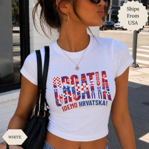 May include: A white short-sleeved t-shirt featuring the word CROATIA in large, bold letters filled with a red and white checkered pattern and blue accents. Below the main text, the phrase IDEMO HRVATSKA! is printed in red and blue lettering.