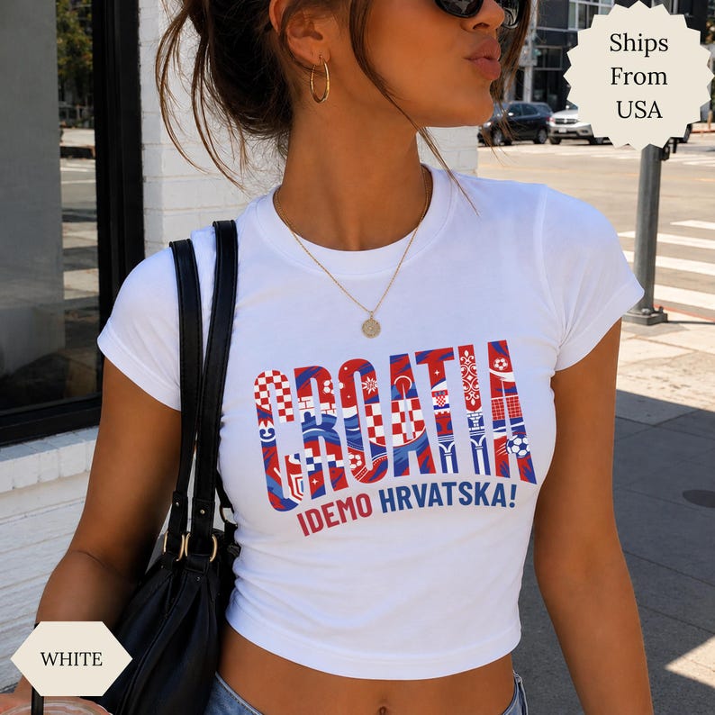 May include: A white short-sleeved t-shirt featuring the word CROATIA in large, bold letters filled with a red and white checkered pattern and blue accents. Below the main text, the phrase IDEMO HRVATSKA! is printed in red and blue lettering.
