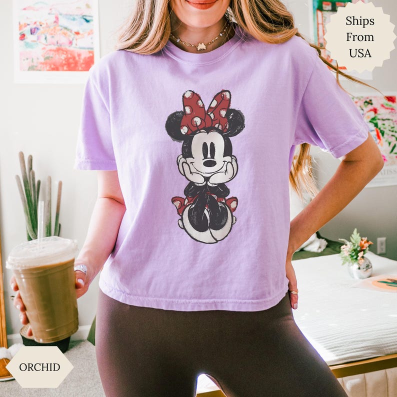 May include: A light purple t-shirt featuring a distressed Minnie Mouse graphic. Minnie Mouse is depicted with a red polka dot bow, black ears, and a red bow on her shoes. The shirt is a relaxed fit.
