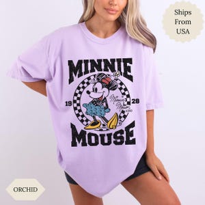 May include: Lilac t-shirt with a distressed Minnie Mouse graphic. The design features the text "MINNIE MOUSE" in black, with Minnie Mouse in the center. The shirt has short sleeves and a loose fit. The colour is described as Orchid.