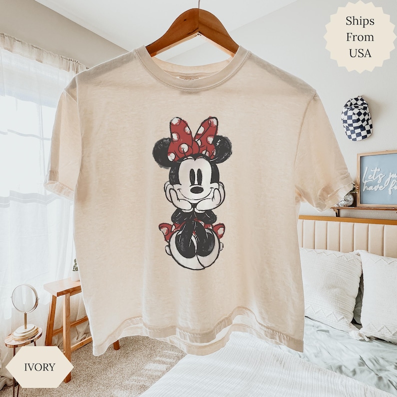 May include: Ivory-colored t-shirt featuring a Minnie Mouse graphic. The graphic depicts Minnie Mouse with a red polka-dot bow and matching accents. The shirt is hanging on a wooden hanger.