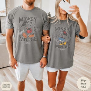 Puede incluir: Dos camisetas de color gris carb&oacute;n con gr&aacute;ficos de estilo vintage. La camiseta de la izquierda muestra a Mickey Mouse con el texto "Mickey Mouse 1928" dentro de un c&iacute;rculo a cuadros. La de la derecha muestra a Minnie Mouse con el texto "Minnie Mouse 1928" dentro de un c&iacute;rculo a cuadros.