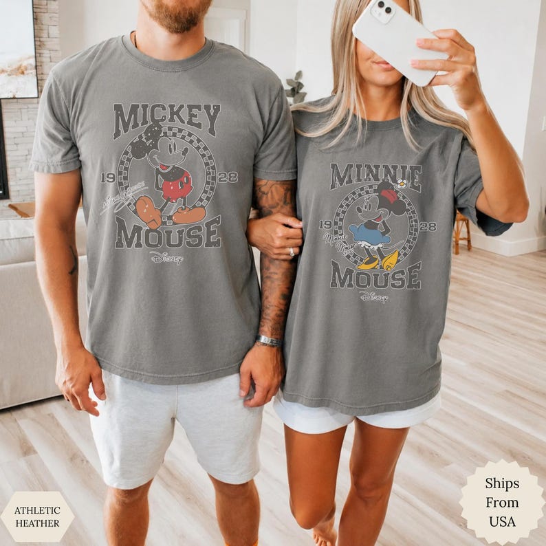 Puede incluir: Dos camisetas de color gris carb&oacute;n con gr&aacute;ficos de estilo vintage. La camiseta de la izquierda muestra a Mickey Mouse con el texto "Mickey Mouse 1928" dentro de un c&iacute;rculo a cuadros. La de la derecha muestra a Minnie Mouse con el texto "Minnie Mouse 1928" dentro de un c&iacute;rculo a cuadros.