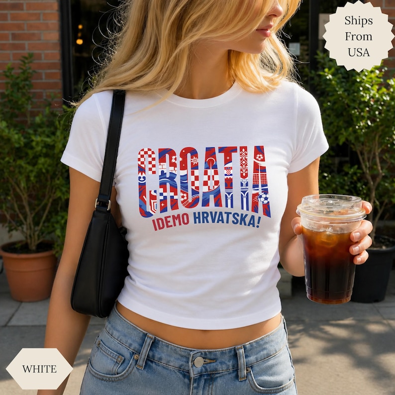 May include: A white short-sleeved t-shirt featuring the word CROATIA in large, stylized red and blue letters with a checkered pattern, and the text IDEMO HRVATSKA! printed in smaller red and blue letters underneath the main graphic design.