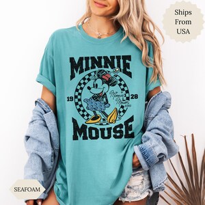 May include: Seafoam green t-shirt with a vintage Minnie Mouse graphic. The design includes the text "MINNIE MOUSE" and the years "1928" and "19". Minnie is shown in a classic pose, wearing a polka-dot skirt and yellow shoes. The shirt is a relaxed fit.