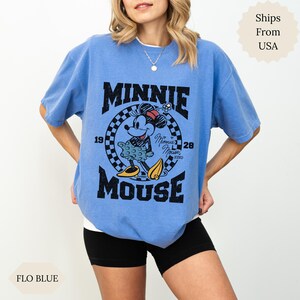 May include: Light blue t-shirt with a Minnie Mouse graphic. The design includes the text "MINNIE MOUSE", the years "1928", and a checkered pattern. The shirt is a 'Flo Blue' colour.