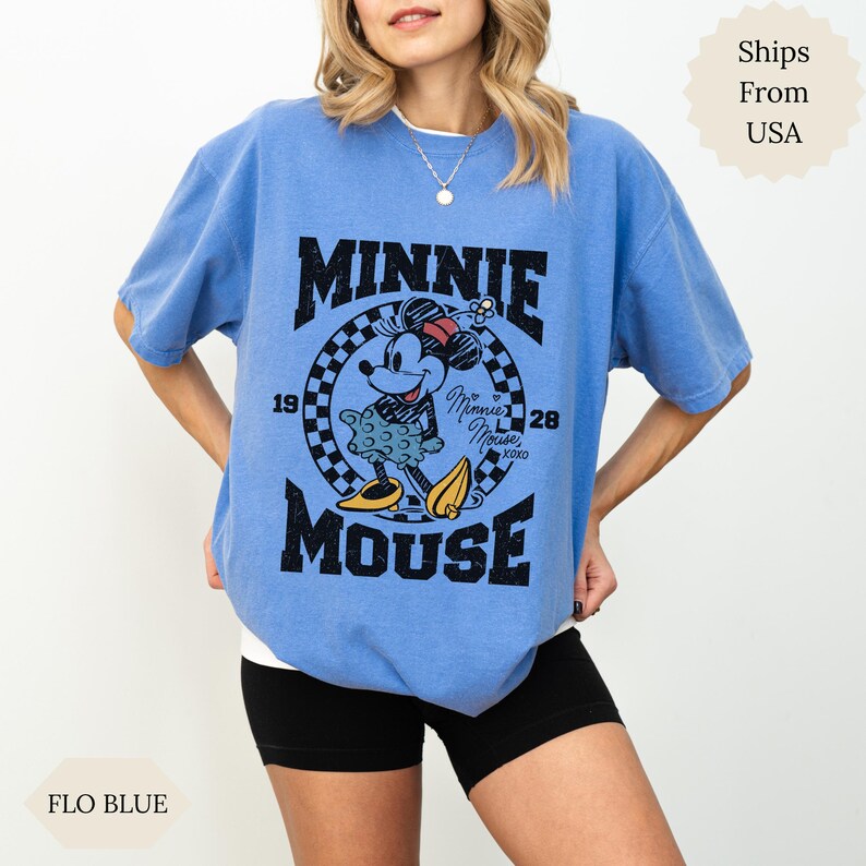 May include: Light blue t-shirt with a Minnie Mouse graphic. The design includes the text "MINNIE MOUSE", the years "1928", and a checkered pattern. The shirt is a 'Flo Blue' colour.