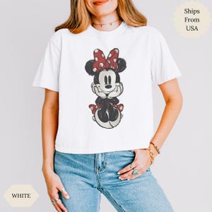 May include: White t-shirt featuring a seated Minnie Mouse graphic. Minnie Mouse is depicted in black, red, and white, with a red polka-dot bow. The shirt is worn by a person wearing blue jeans.