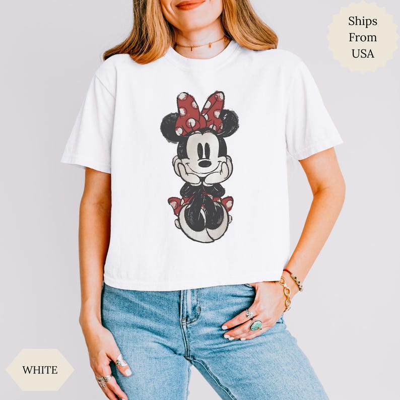 May include: White t-shirt featuring a seated Minnie Mouse graphic. Minnie Mouse is depicted in black, red, and white, with a red polka-dot bow. The shirt is worn by a person wearing blue jeans.