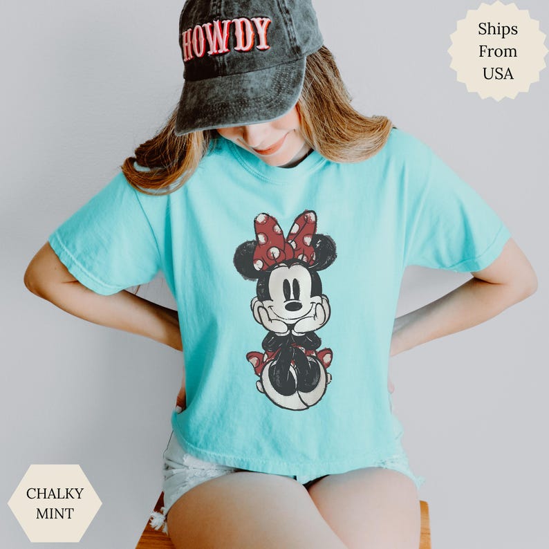 May include: A light blue t-shirt featuring a Minnie Mouse graphic. Minnie is depicted in a seated pose, wearing a red polka dot bow. The shirt is a comfortable, casual style.