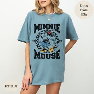 May include: Ice blue t-shirt with a Minnie Mouse graphic. The design includes the text "MINNIE MOUSE" with the years "1928" and "19" on either side of a checkered circle. Minnie Mouse is wearing a red bow and yellow shoes.