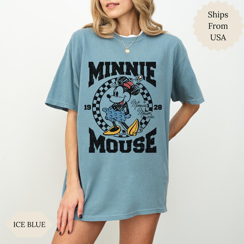 May include: Ice blue t-shirt with a Minnie Mouse graphic. The design includes the text "MINNIE MOUSE" with the years "1928" and "19" on either side of a checkered circle. Minnie Mouse is wearing a red bow and yellow shoes.