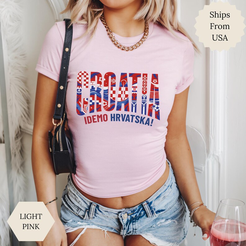 May include: A light pink short-sleeve t-shirt featuring the word CROATIA in large block letters filled with red, white, and blue patterns, and the text IDEMO HRVATSKA! printed in blue below. The shirt has a crew neckline and a fitted silhouette.