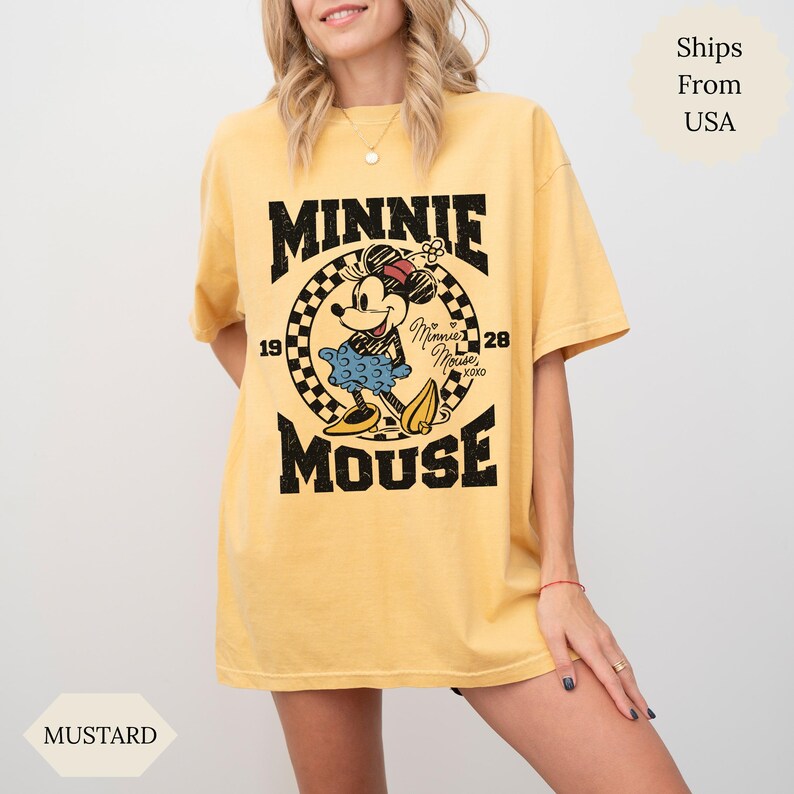 May include: Mustard-coloured t-shirt with a Minnie Mouse graphic. The design features the words "MINNIE MOUSE" in bold black letters, a chequered circle, and the year "1928". Minnie is shown in her iconic outfit.