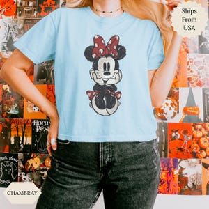 May include: Light blue t-shirt featuring a distressed graphic of Minnie Mouse with a red polka dot bow. The shirt is made of soft material and is a casual wear item. The word "CHAMBRAY" is printed on the bottom left.