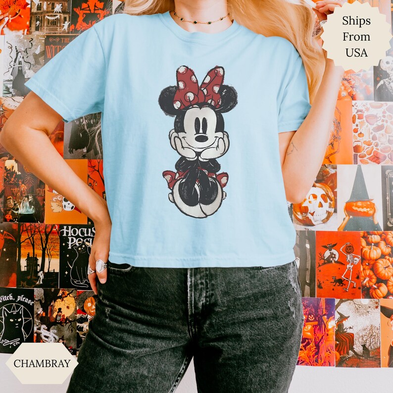 May include: Light blue t-shirt featuring a distressed graphic of Minnie Mouse with a red polka dot bow. The shirt is made of soft material and is a casual wear item. The word "CHAMBRAY" is printed on the bottom left.