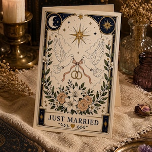Op de afbeelding: Een trouwkaart met een hemels ontwerp. Twee duiven houden een lint boven een bloemstuk. De kaart heeft de tekst "JUST MARRIED" in een banner met een hart en bladeren.
