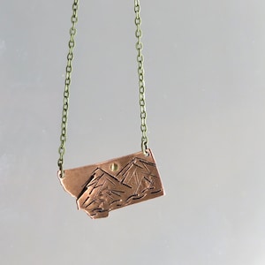 May include: A copper necklace with a pendant shaped like the state of Montana. The pendant features an engraved mountain range and a small sun.