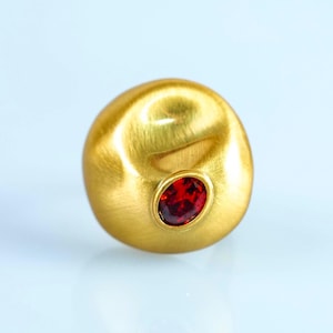 Könnte beinhalten: Ein Ring mit einer großen, gebürsteten goldfarbenen Kuppel in unregelmäßiger, organischer Form. Ein einzelner, kleiner, oval geschliffener, tiefroter Edelstein ist mittig in die Goldoberfläche eingelassen und bildet einen Kontrast.