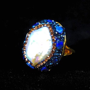 May include: A gold-toned ring featuring a large, irregular white baroque pearl at the centre. The pearl is surrounded by a border of small, shimmering copper-coloured crystals, followed by an outer layer of deep blue crystals and blue stone accents.