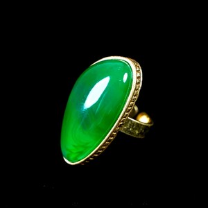 May include: A ring featuring a large, smooth, oval-shaped green stone set in a gold-toned metal band. The setting has a textured, patterned edge surrounding the vibrant green cabochon, which displays subtle internal swirls and a reflective surface.