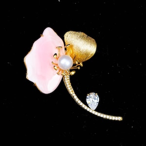 May include: A floral brooch featuring a smooth, pale pink petal, a textured gold-tone leaf, and a central white pearl. The curved stem is encrusted with small clear stones and features a single pear-shaped clear stone accent near the base.