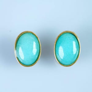 May include: A pair of oval-shaped stud earrings featuring smooth, polished turquoise-colored cabochons set within simple gold-toned metal frames. The earrings have a glossy finish and a uniform, vibrant blue-green appearance on a light background.