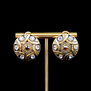 May include: A pair of round gold-tone earrings featuring a textured, interwoven metal pattern. Each earring is accented with a central deep red stone and several small, clear, sparkling crystals embedded throughout the circular design.