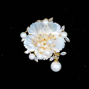 Op de afbeelding: Een goudkleurige broche in de vorm van een bloem met witte parelmoeren bloemblaadjes. Het midden is versierd met meerdere ronde witte parels en kleine heldere baguette-geslepen kristallen. Extra parels en marquise-geslepen kristallen steken uit.