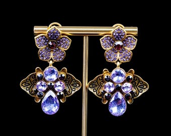 May include: A pair of gold-toned earrings featuring a flower-shaped top encrusted with small purple crystals and a central dark red stone. The lower section displays a fan-like metal frame with light purple crystals and a large teardrop-shaped stone.