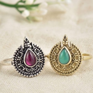 May include: Two rings featuring ornate circular settings with teardrop-shaped stones. The left ring is silver-toned with a deep red stone, while the right ring is gold-toned with a turquoise-colored stone. Both have intricate metalwork details.