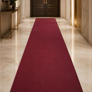 May include: A long, rectangular runner rug in a solid deep burgundy red color. The rug features a smooth, uniform texture across its entire surface and is finished with a clean, matching bound edge that runs along the perimeter of the piece.