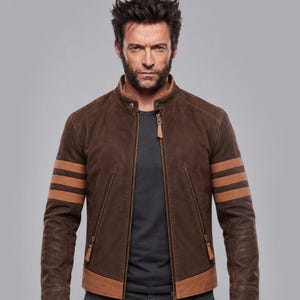Könnte beinhalten: Eine dunkelbraune Wildlederjacke mit Stehkragen, durchgehendem Frontreißverschluss und drei horizontalen hellbraunen Streifen an jedem Ärmel. Die Jacke verfügt über zwei schräge Reißverschlusstaschen auf der Vorderseite und einen hellbraunen Saum.