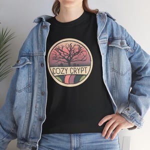 May include: Black t-shirt featuring a circular graphic with a bare tree silhouette against a sunset sky. The graphic includes the text "COZY CRYPT" in a curved font, with a colorful striped design at the bottom. The shirt is worn with a denim jacket.