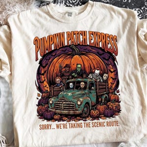 May include: A cream-colored t-shirt featuring a graphic of a teal vintage truck carrying horror movie characters in front of a large orange pumpkin. The text reads PUMPKIN PATCH EXPRESS and SORRY... WE'RE TAKING THE SCENIC ROUTE. Orange pumpkins surround the truck.