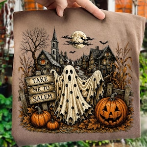 May include: A brown t-shirt featuring a graphic of a polka-dot ghost standing before a wooden sign that reads TAKE ME TO SALEM. The scene includes a full moon, bats, a dark village with a church, corn stalks, and two orange jack-o'-lanterns.