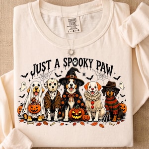 May include: A cream-colored long-sleeve shirt featuring a graphic of five dogs in Halloween costumes with the text "JUST A SPOOKY PAW". The dogs are dressed as a ghost, a hockey-masked figure, a witch, a clown, and a striped-sweater character.