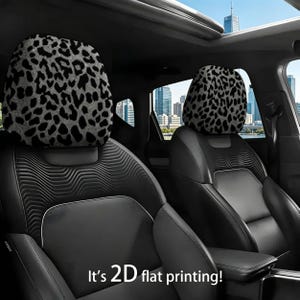 Op de afbeelding: Twee hoofdsteunen voor auto's met een zwart-grijs luipaardprintpatroon. De hoofdsteunen zijn geïnstalleerd in een auto met zwarte leren stoelen. De tekst "It's 2D flat printing!" staat onderaan de afbeelding.