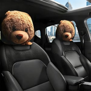 May include: Two car headrest covers shaped like teddy bear heads. The plush bears are brown with tan snouts and dark eyes. The headrests are attached to black car seats with a city skyline visible through the car windows.