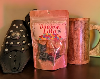 May include: A clear bag of gummy sweets labelled "Dungeon Loot" with a colourful graphic. Beside it is a wooden mug and a black leather gauntlet with silver studs and a decorative emblem.