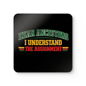 May include: Square black coaster with the text "DEAR ANCESTORS I UNDERSTAND THE ASSIGNMENT" in green, yellow, and red. The coaster has rounded corners and a glossy finish. The text is in a bold, stylized font.