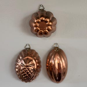 Könnte beinhalten: Drei kupferfarbene Vintage-Formen zur Herstellung von Desserts. Die Formen umfassen eine Blumenform, ein Oval mit Ananas-Design und ein geripptes Oval. Jede Form hat eine kleine Metallschlaufe zum Aufhängen.